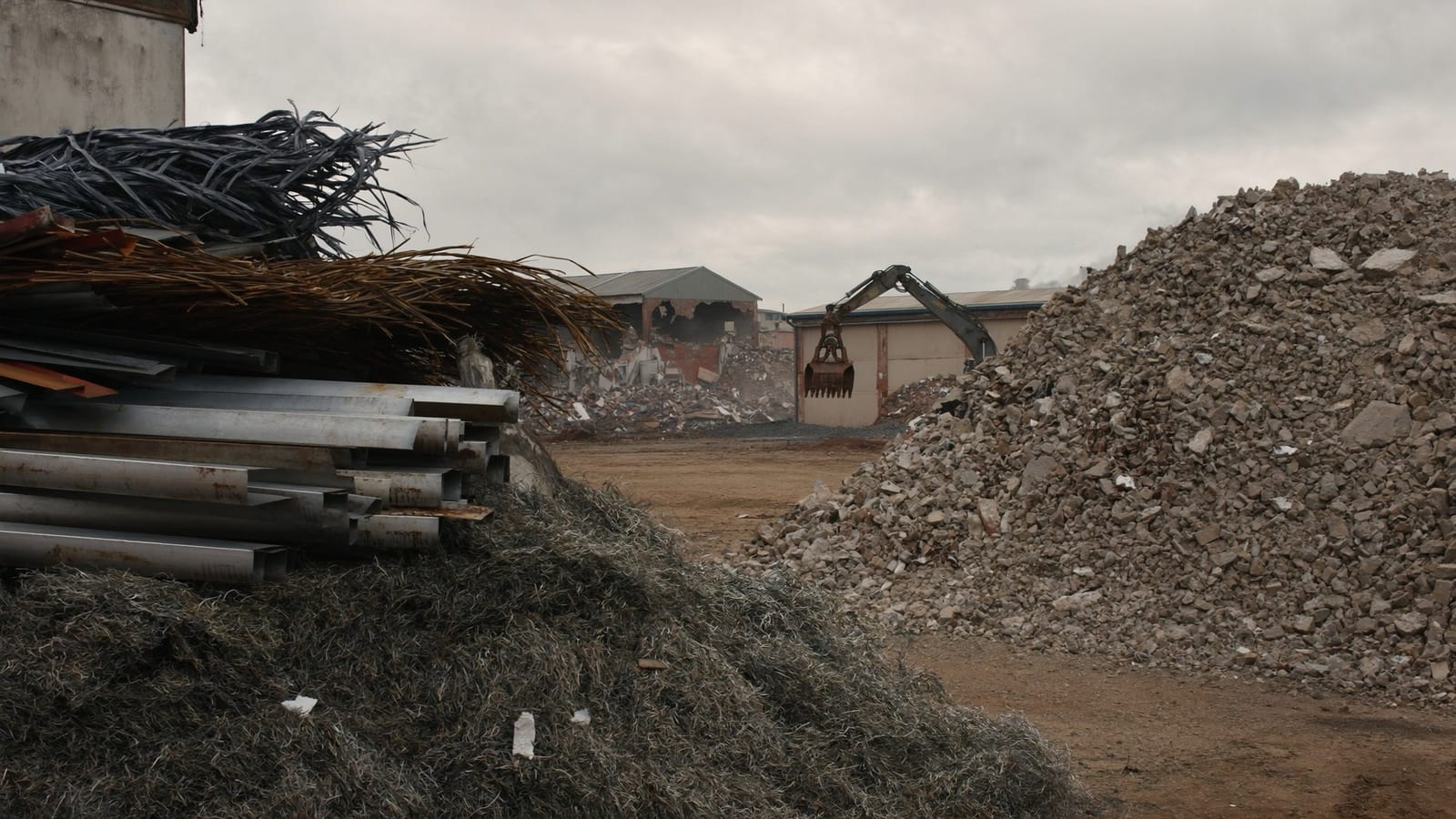 Separated stockpiles of scrap steel and crushed concrete on a demolition site with an excavator and sorting grapple behind