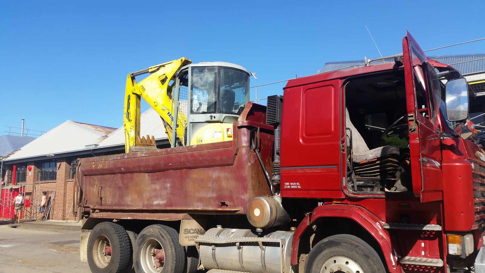 Red tipper truck carrying a yellow mini excavator in its body on a job site