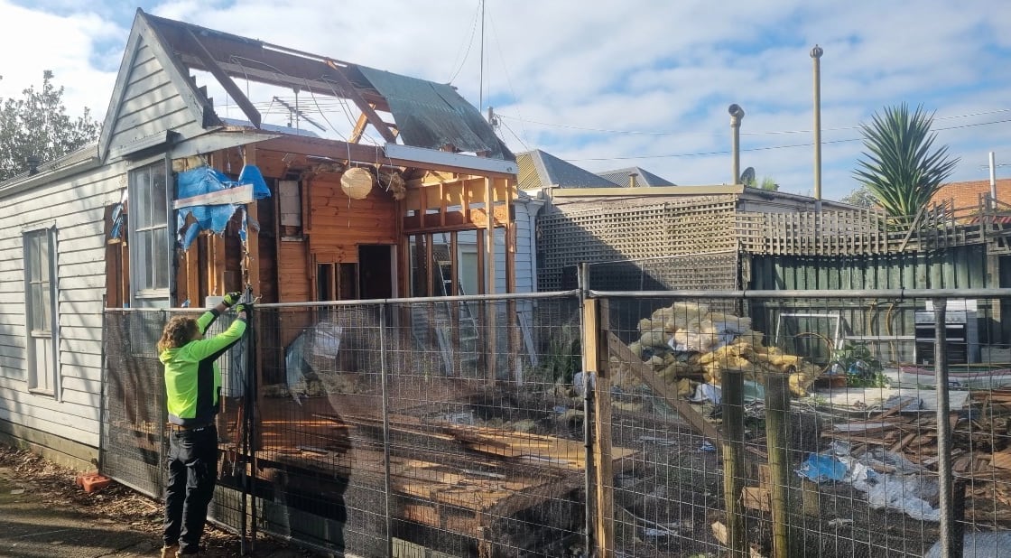 Worker in high-visibility clothing securing temporary mesh fencing across the front of a weatherboard house with its roof partly stripped