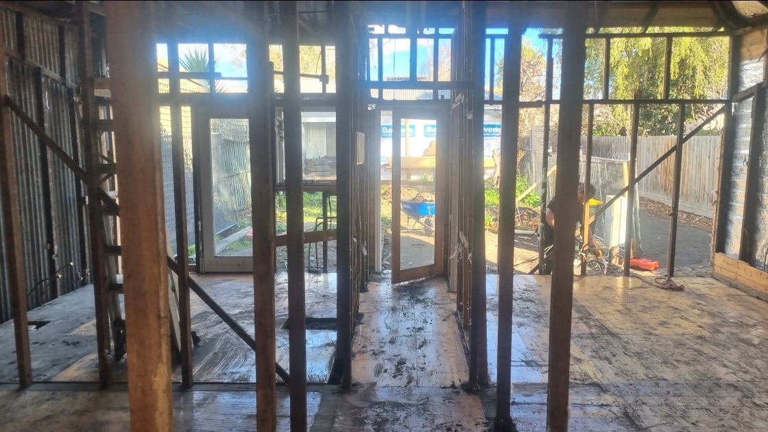 Stripped house interior looking through bare door openings to the back yard, original floorboards and wall framing exposed