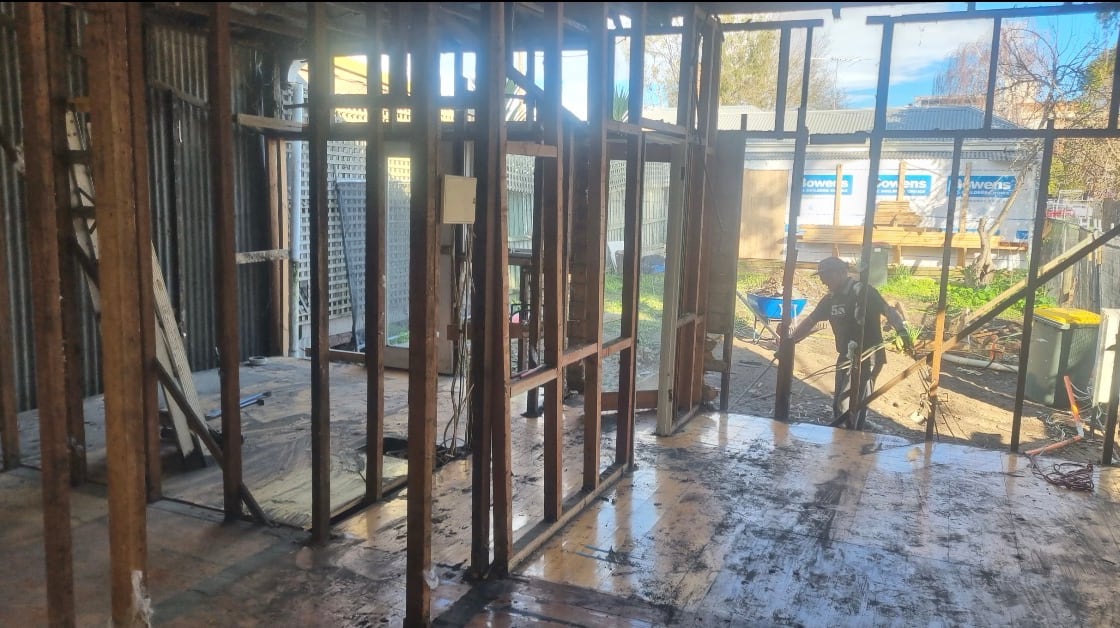 Interior of a weatherboard house stripped to bare timber studs and corrugated iron sheeting, with a worker loading a wheelbarrow in the yard beyond