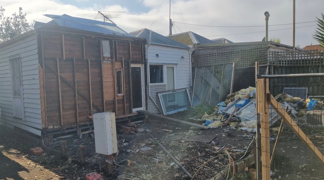 Removed window frames stacked in a back yard beside piles of demolition debris and insulation, with the stripped house behind